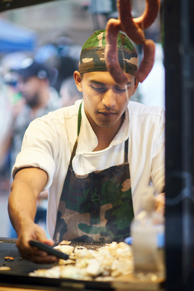 Focused man in uniform cooking food on iron tray surrounded with smoke at food market